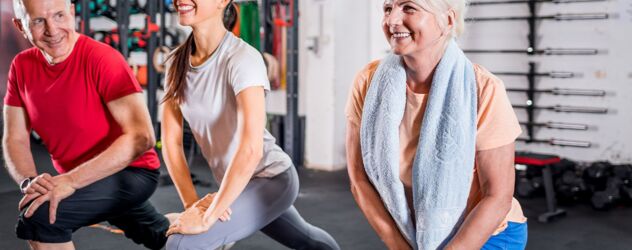 In einem Fitnessstudio absolviert eine Fitnesstrainerin mit zwei Senioren eine Übung. Sie stützen ihre Arme auf das linke Knie, während sie mit dem rechten Bein einen Ausfallschritt machen