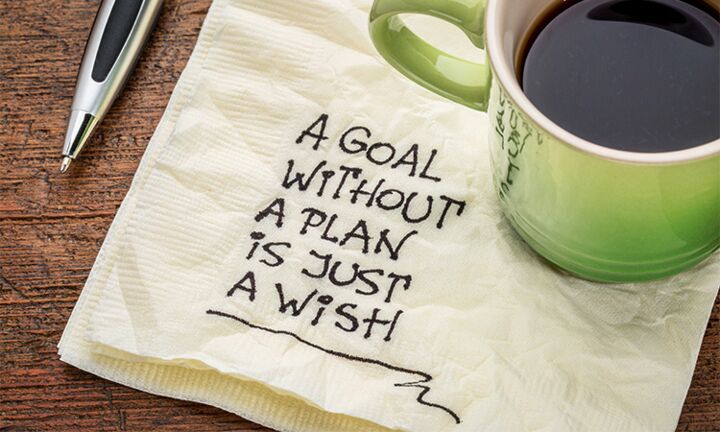 Auf einem Tisch steht eine Tasse Kaffee auf einer Serviette mit der Inschrift "A goal without a plan is just a wish". Daneben liegt ein Kugelschreiber.