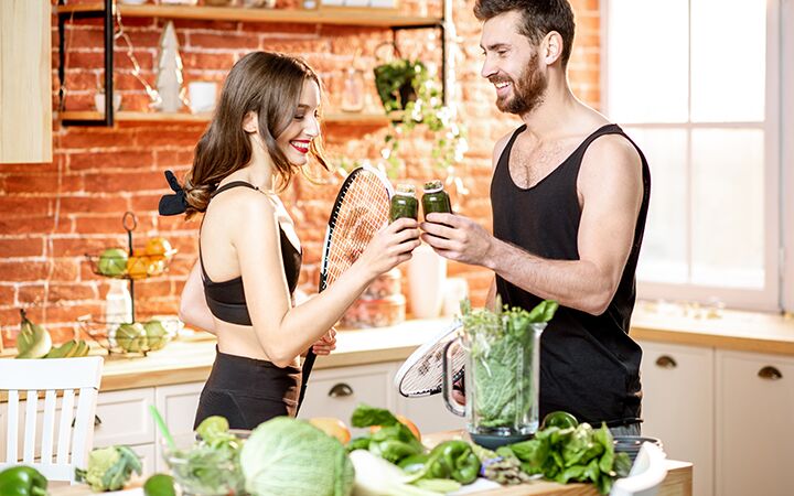 Junges sportliches Paar, das einen vegetarischen Snack zu sich nimmt und einen Smoothie trinkt, während es mit Schlägern in der Küche zu Hause steht