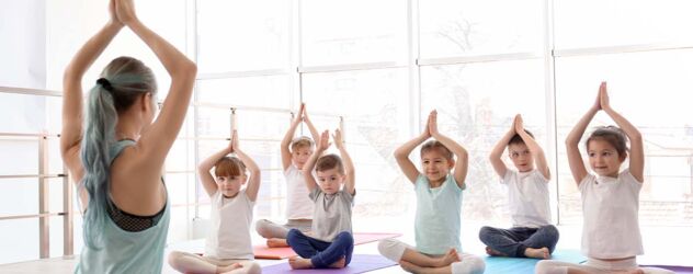 In einem hellen Sportraum sitzt eine junge Yogalehrerin im Schneidersitz und hält die Arme über ihrem Kopf zusammen. Vor sich sitzen einige Kinder auf Yogamatten und machen die Übung nach.