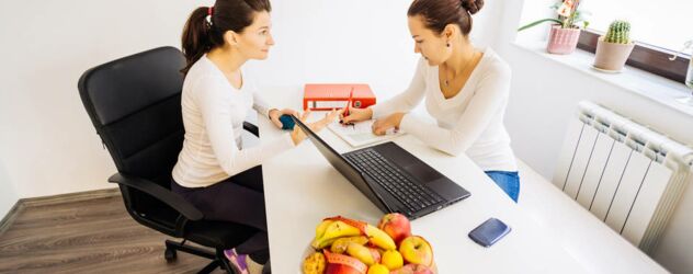Zwei Frauen sitzen an einem Schreibtisch mit Obst und einem Laptop.