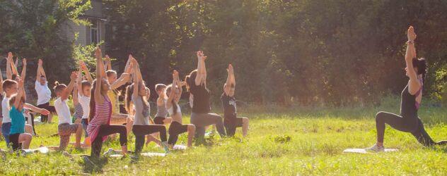 Auf einer Wiese macht eine Yogalehrerin eine Yogaübung vor. Eine große Menge an Kindern macht ihr diese nach. Sie strecken dabei die Arme in die Luft und stehen halb aufrecht auf unterschiedlichen Yogamatten.
