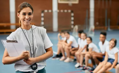 Eine junge Sportlehrerin steht in der Sporthalle von der Gruppe und lächelt.