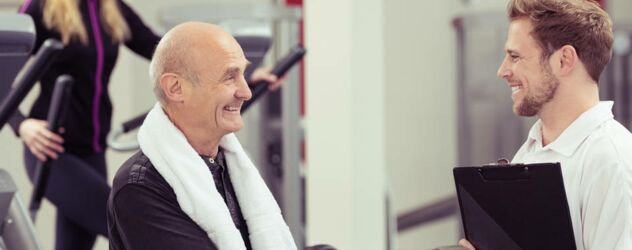 Lächelnder älterer Mann im Gespräch mit jungem Fitnesstrainer im Fitnessstudio.