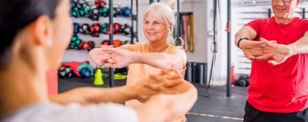 In einem Fitnessstudio führt eine junge Seniorentrainerin gerade mit zwei älteren Menschen ein Seniorentraining durch. Dabei stehen diese breitbeinig da und drücken beide Handflächen von ihrem Körper weg