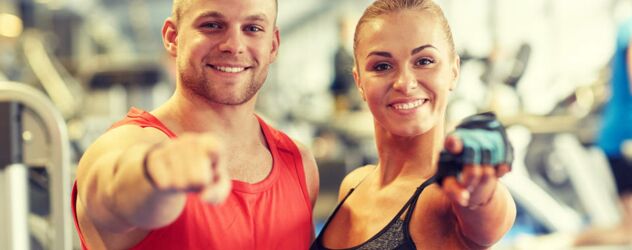 ein Mann und eine Frau zeigen in die Kamera und grinsen im Fitnessstudio