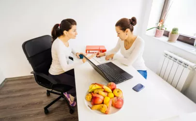 Zwei Frauen sitzen an einem Schreibtisch mit Obst und einem Laptop.