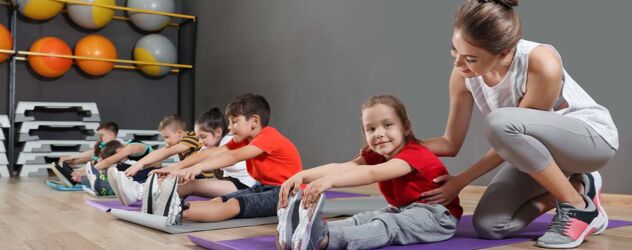 Junge Yogalehrerin, die gerade dabei ist, mehrere Kinder, sitzend auf verschiedenen Yogamatten, zu betreuen. Die Kinder sitzen dabei mit ausgestreckten Beinen und versuchen die Finger bis zu den Zehenspitzen zu strecken.