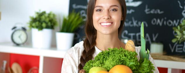 eine junge Frau mit einer Einkaufstüte, voller Lebensmittel, schaut lächelnd in die Kamera