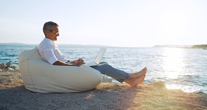 Resilienter Mann arbeitet im Urlaub am Strand mit Laptop im Sitzsack mit Meerblick.